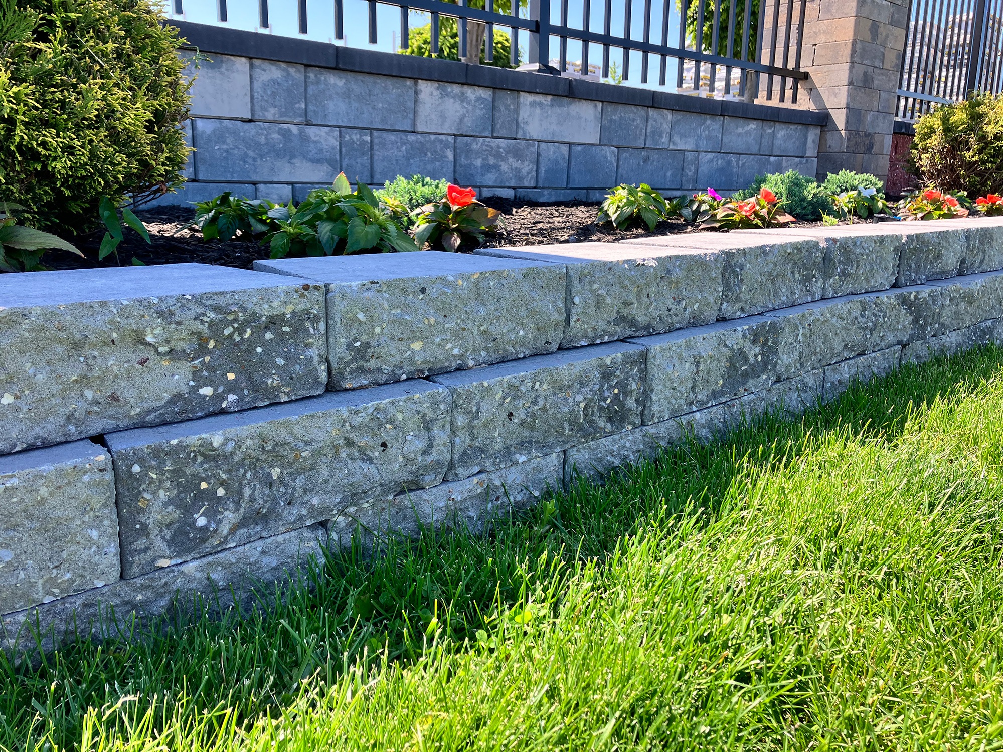 Stone wall garden with tiered blocks, lush green grass, and colorful flowers planted along the upper ledge.
